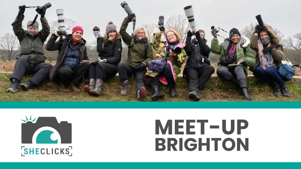 Group of women photographers holding cameras outdoors; banner below reads “SheClicks Meet-Up Brighton” with camera logo.