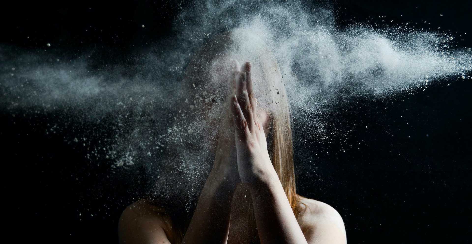 Person with hands clapped together in front of their face as a cloud of white powder explodes outward against a dark background.