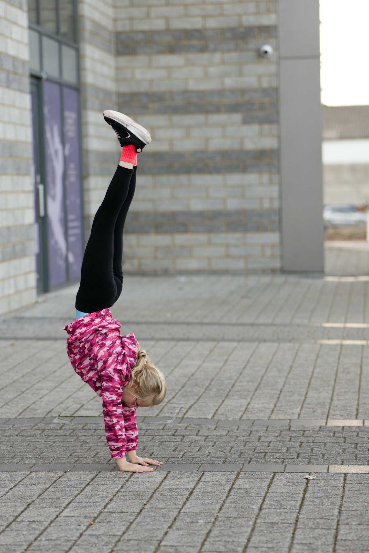 Young child in a pink patterned jacket doing a handstand on a paved area outdoors.