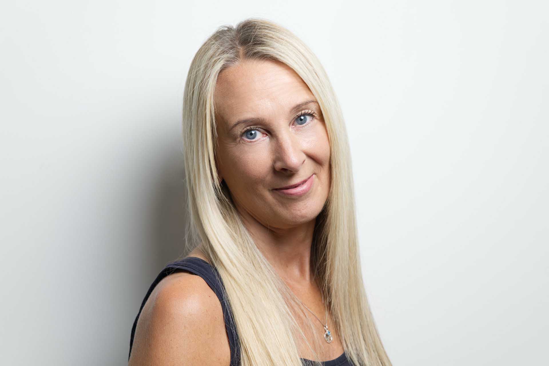 Studio portrait of a blonde woman with long straight hair wearing a sleeveless top, photographed against a light background.