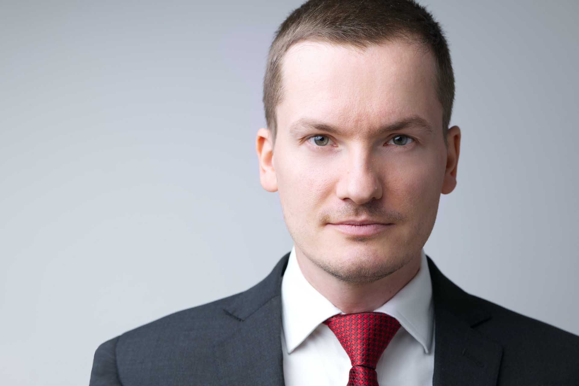 Head-and-shoulders portrait of a man wearing a dark suit, white shirt, and red tie against a light gray background.