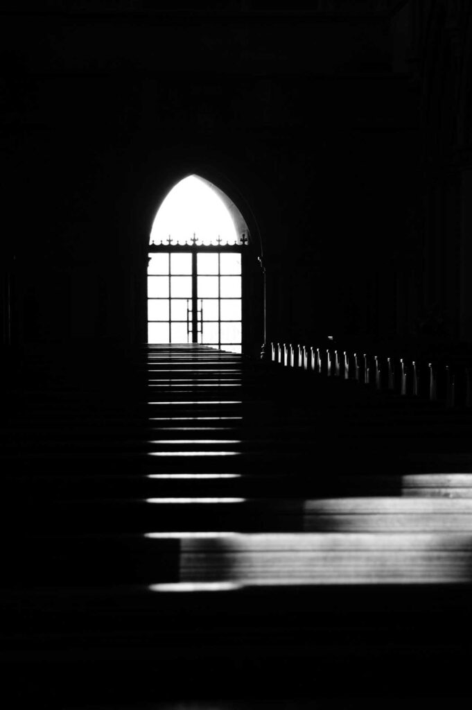 High-contrast black-and-white view down a dark church aisle toward a bright arched doorway, with pews fading into shadow.