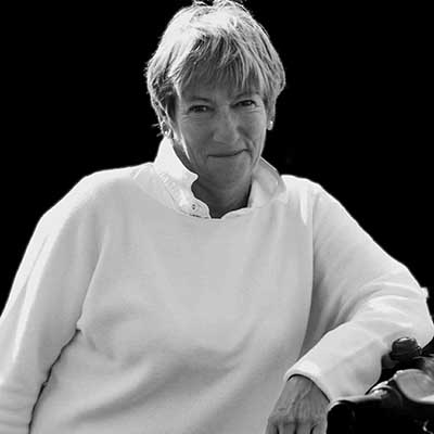 Black-and-white portrait of Beverley Birchley wearing a white sweater, short light hair, and a gentle smile, leaning on a railing against a dark background.