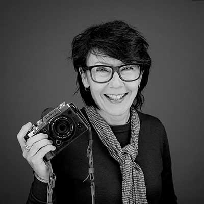 Black-and-white studio portrait of Jayne Odell smiling with short dark hair and glasses, holding a mirrorless camera, wearing a dark top and patterned scarf against a plain background.