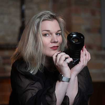 Woman (Tigz Rice) with shoulder-length blonde hair holds a camera close to her face, looking directly toward the viewer, wearing a dark top and watch, posed indoors against a softly blurred brick wall.