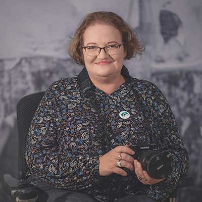 Smiling woman (Sarah Williams) with short light brown hair and glasses sits in a chair holding a camera, wearing a patterned blouse with a round pin, posed indoors against a muted photographic backdrop.