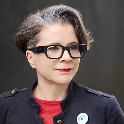 Short-haired person (Angela Nicholson) with gray-streaked dark hair wearing black rectangular glasses, a navy jacket over a red top, and a small round pin, looking slightly to the side against a dark background.