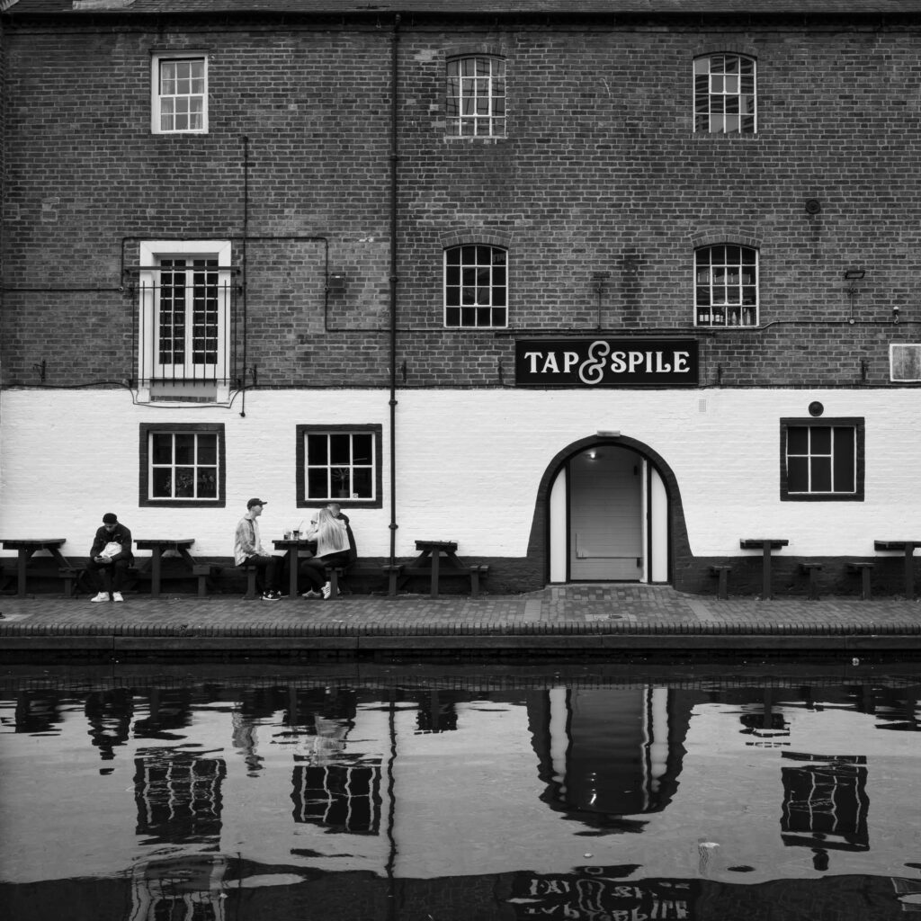 Brick facade of the Tap & Spile pub in Birmingham, with people seated outside and reflections in the canal.