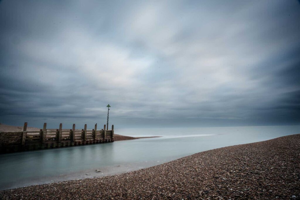 Wide view of a pebble beach and groyne under heavy clouds, long-exposure water forming a pale channel to the sea.