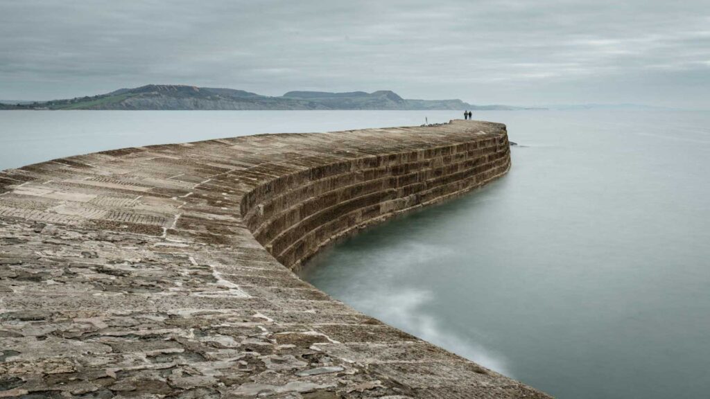 Color version of a curved stone pier extending into pale green water under an overcast sky.