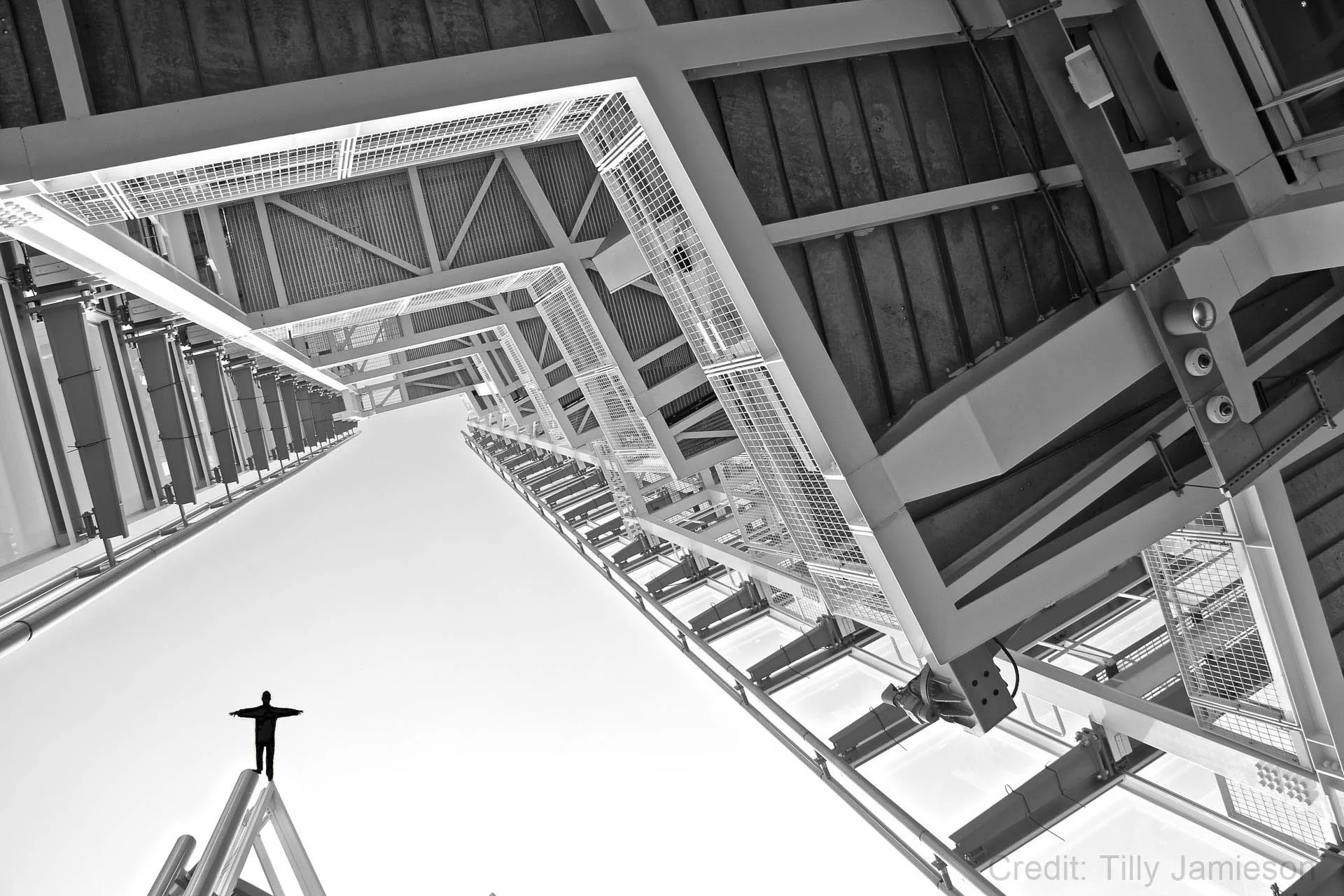 Person standing on a high structure with arms outstretched, surrounded by geometric steel architecture seen from below in black and white.