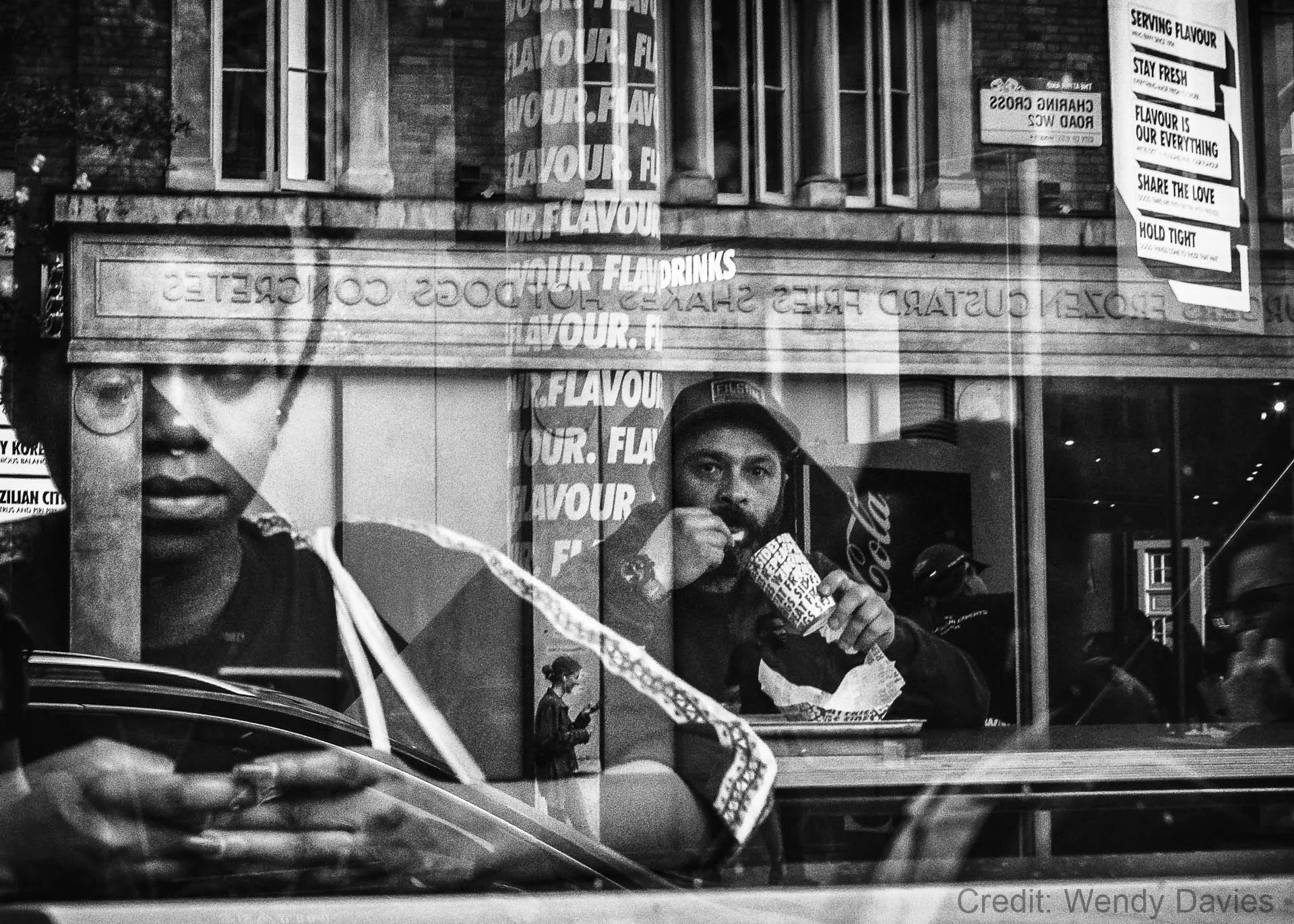 Black and white image of a reflection in a window showing a man eating inside a restaurant. The reflection also includes a woman holding a phone, building facades, and text signs saying 'FLAVOUR' and other phrases. The scene is layered with reflectio