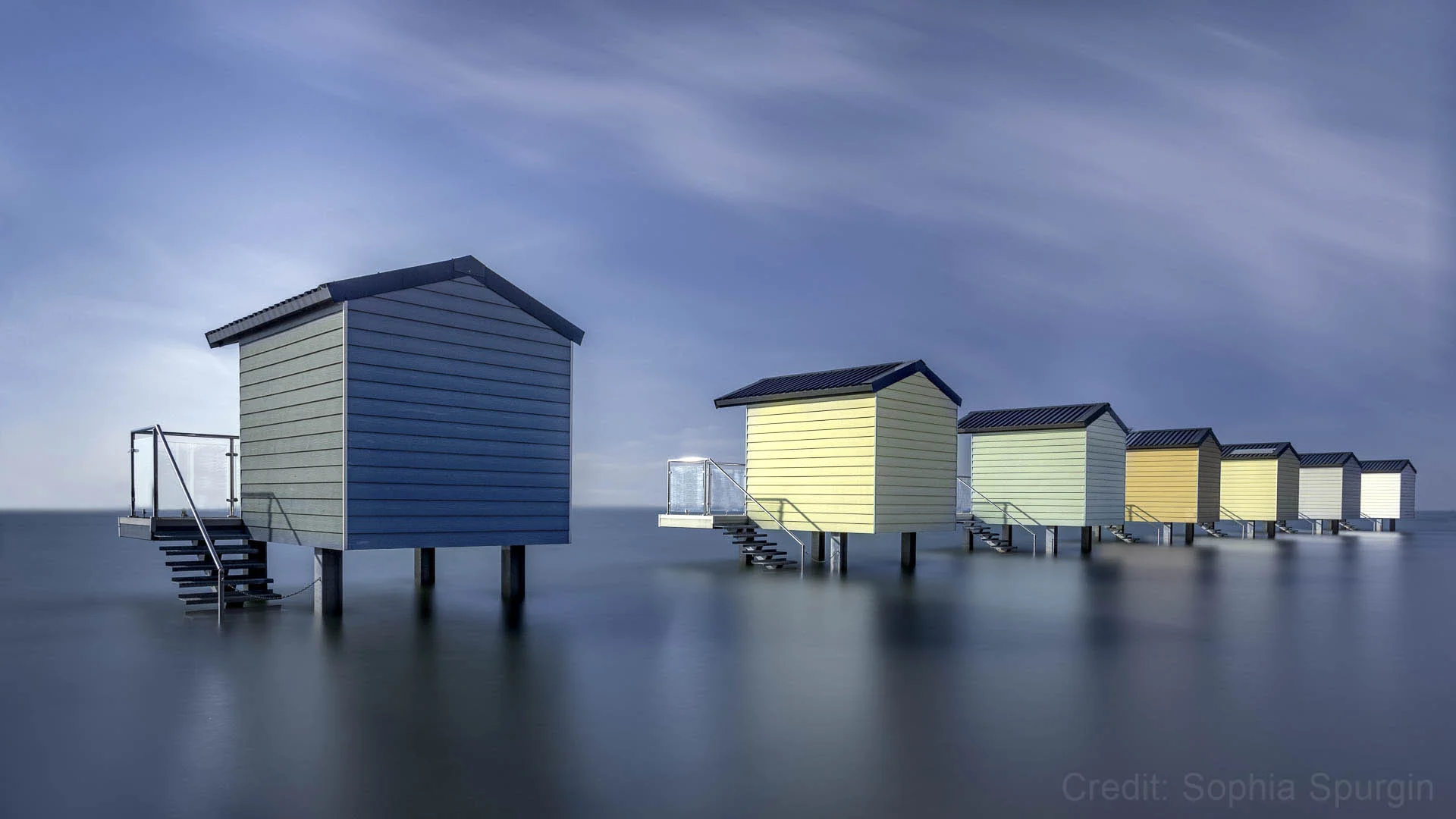 Row of colorful beach huts on stilts over calm water against a cloudy sky.