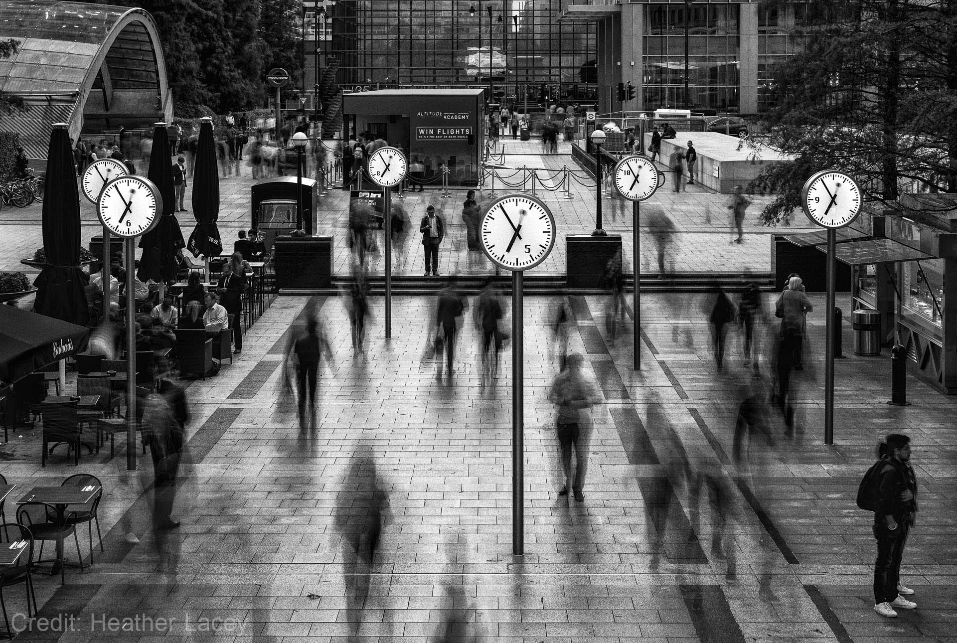Blurred pedestrians walking in a city square with large clocks on poles, creating a dynamic, time-lapse effect.