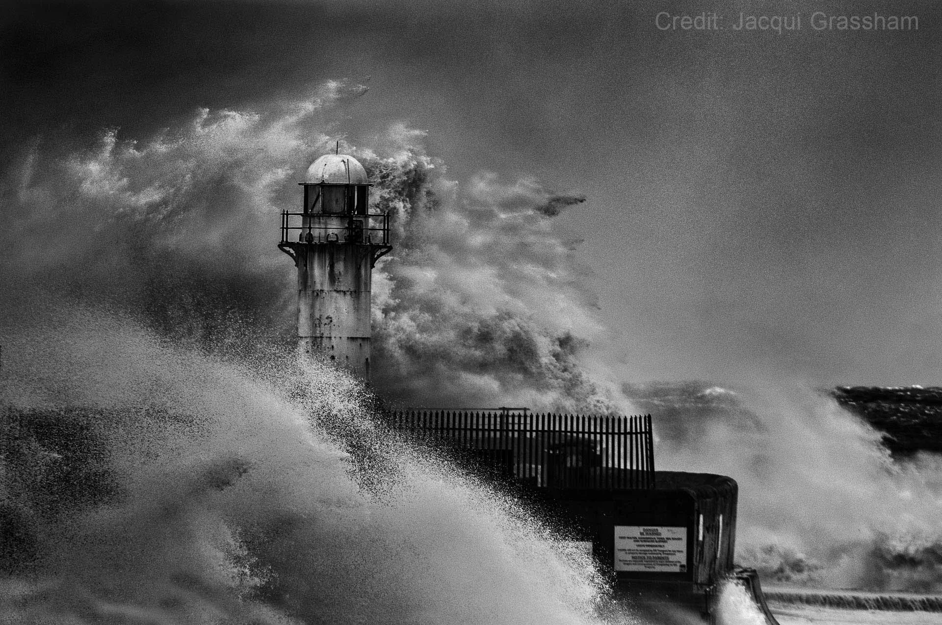 Black and white photo of a lighthouse being hit by large waves during a storm.