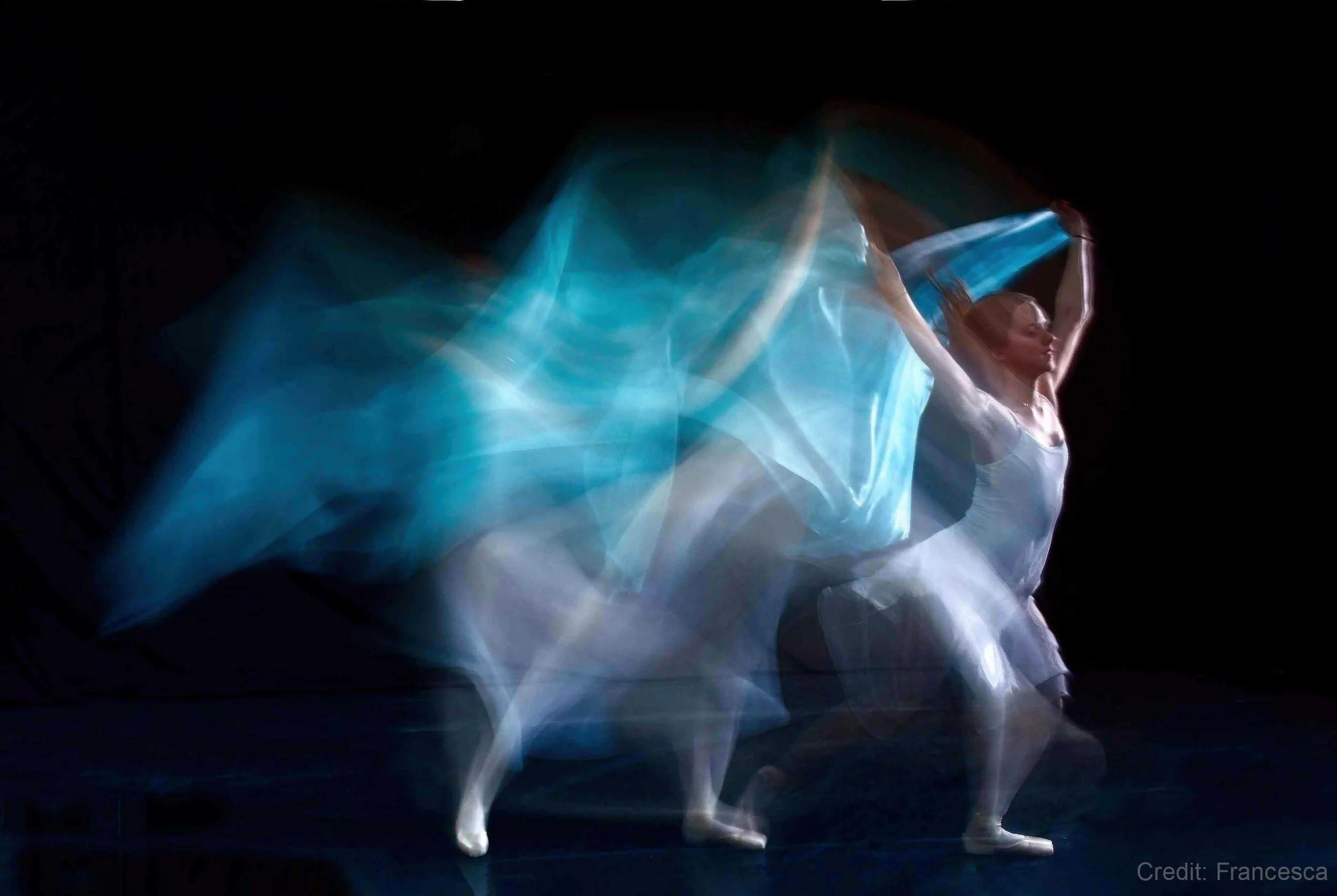 Blurred motion image of a ballet dancer with flowing blue fabric on a dark background.