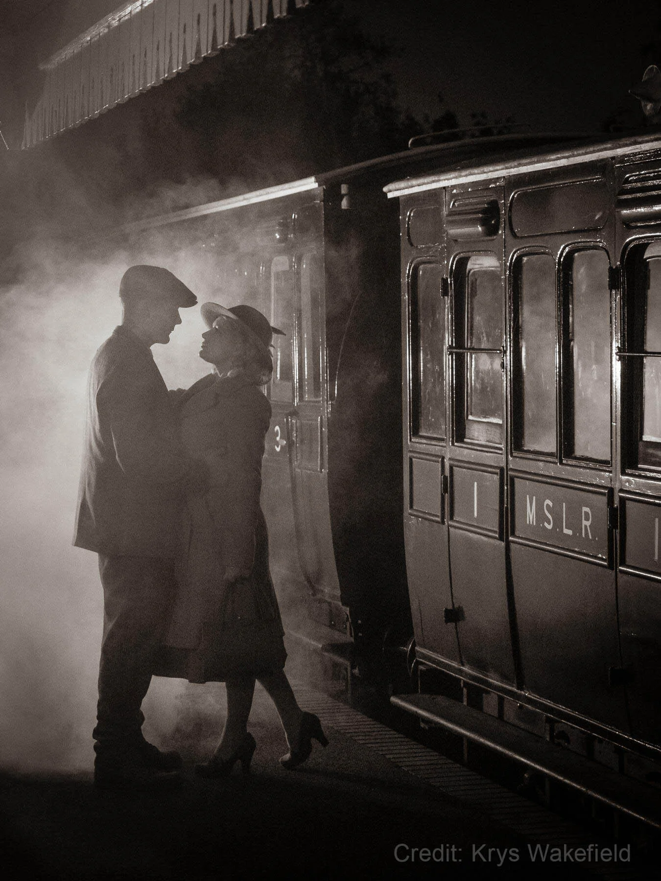 Vintage-style black and white photo of a couple next to an old-fashioned train, with mist and dramatic lighting.