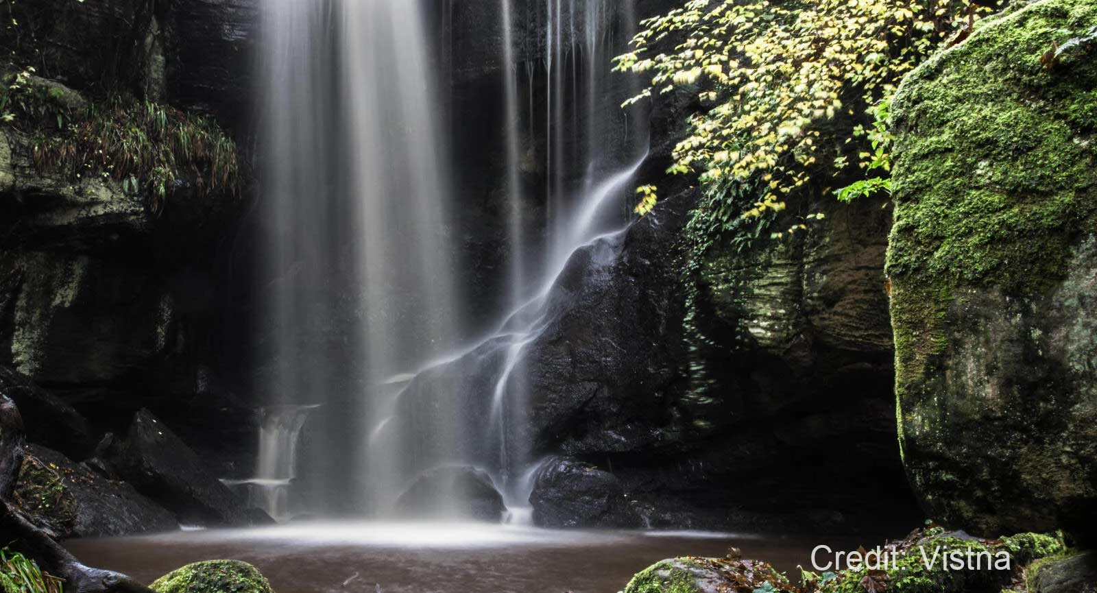 Waterfall cascading into a pool surrounded by moss-covered rocks and lush vegetation.