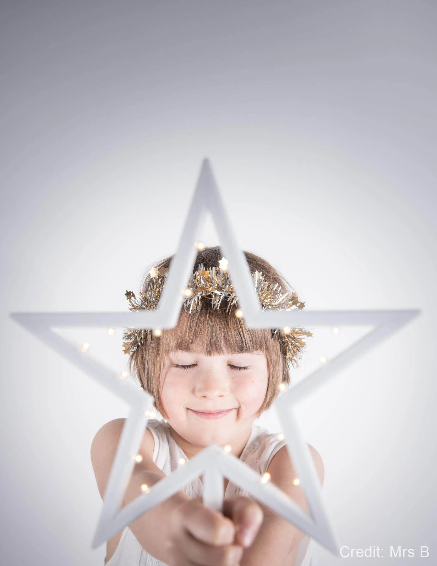 Child holding a lit star decoration, smiling with closed eyes, wearing a tinsel headband.