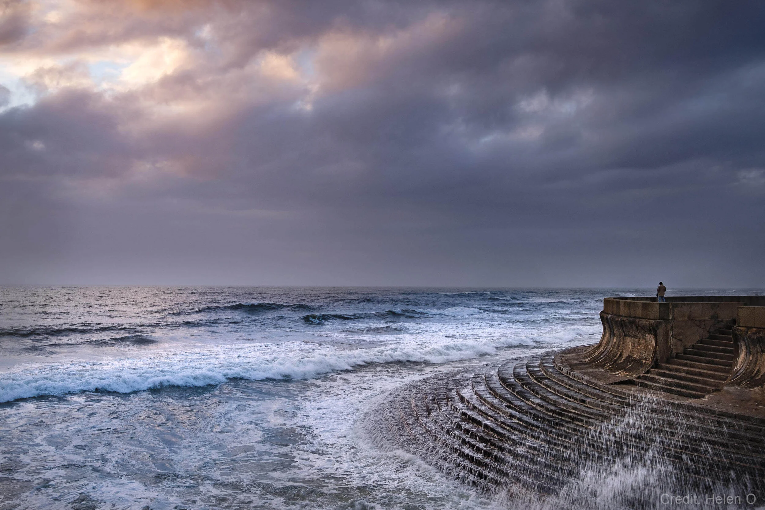 Coastal scene with ocean waves crashing against a curved concrete seawall, person standing on the edge, and dramatic cloudy sky.