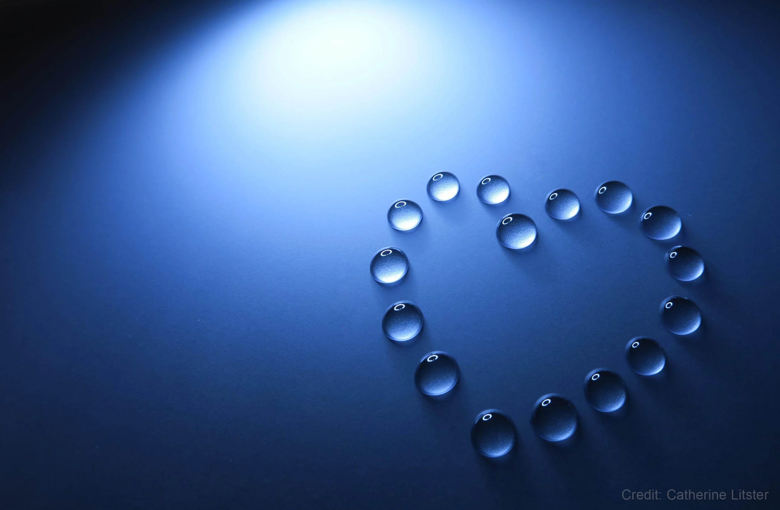 Water droplets arranged in a heart shape on a blue background, illuminated by a soft light from the top left corner.