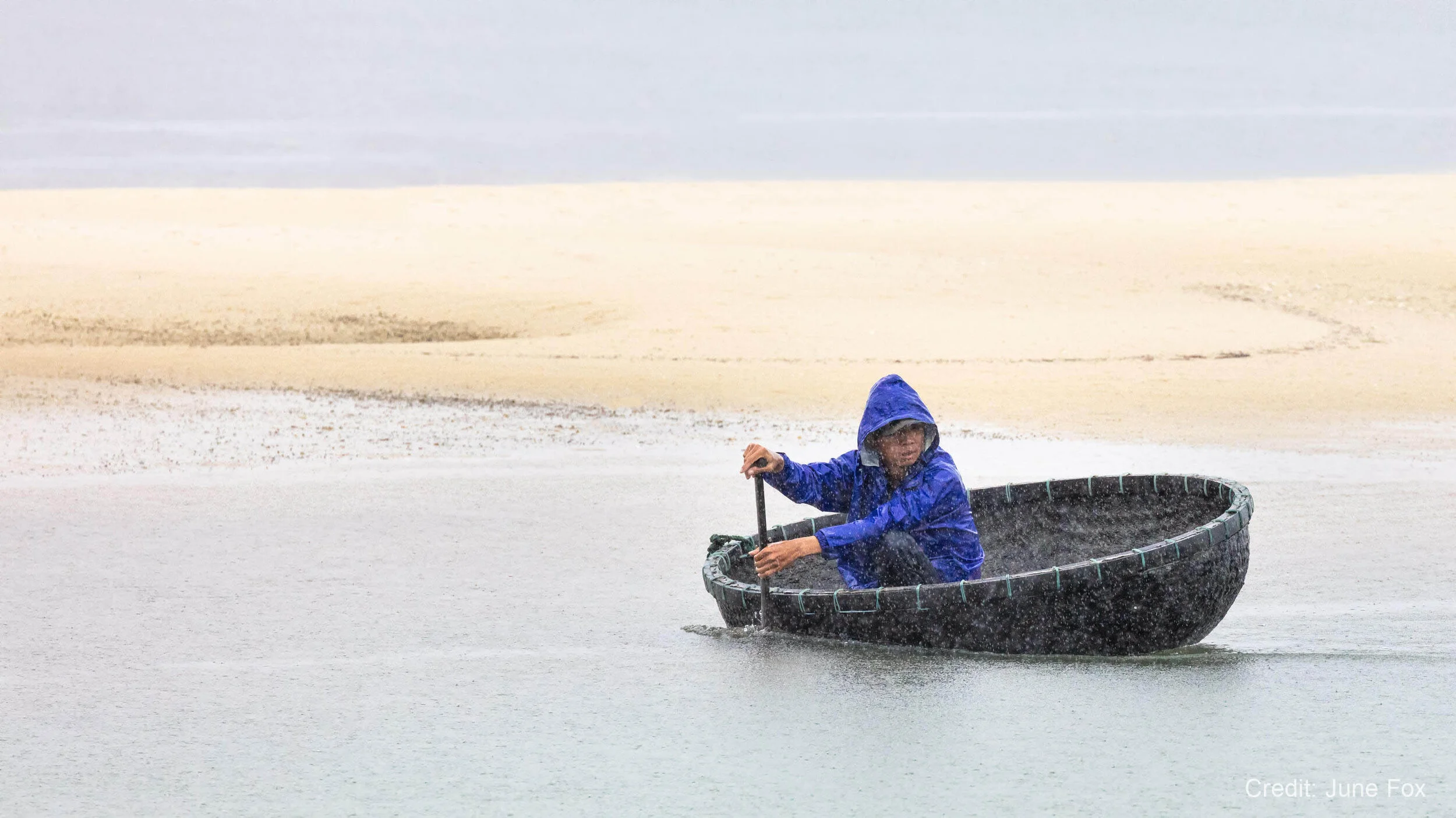 Person in a blue raincoat rowing a black circular boat on a rainy day, with sandy shore in the background.