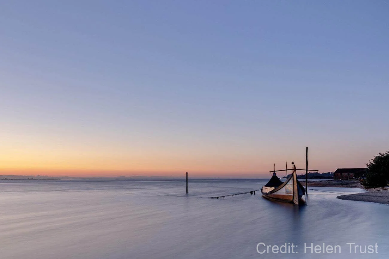 Calm seascape with a wooden boat anchored near the shore at sunset, featuring a gradient sky from orange to blue and gentle waves lapping the beach.