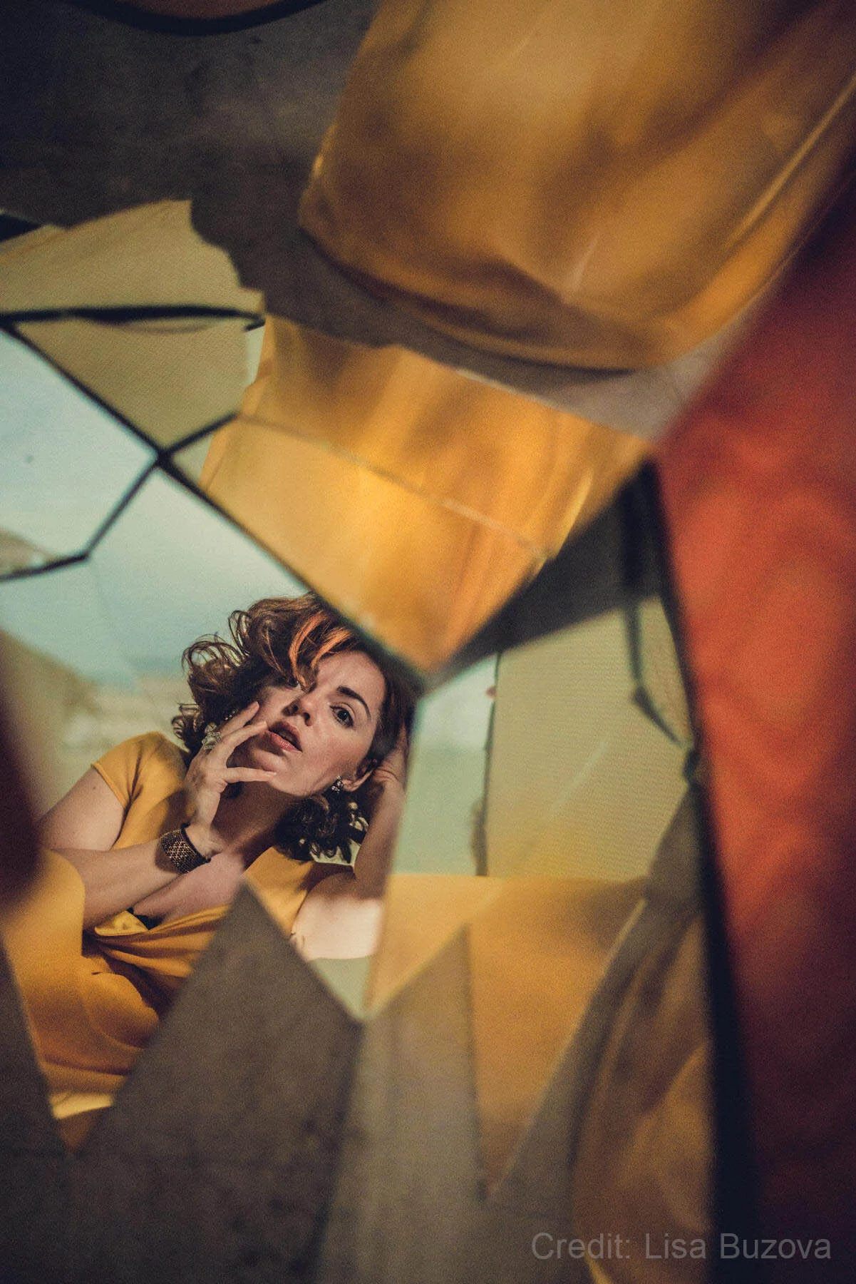 A woman in a yellow dress reflected in a broken mirror, looking at the camera with her hand on her face.