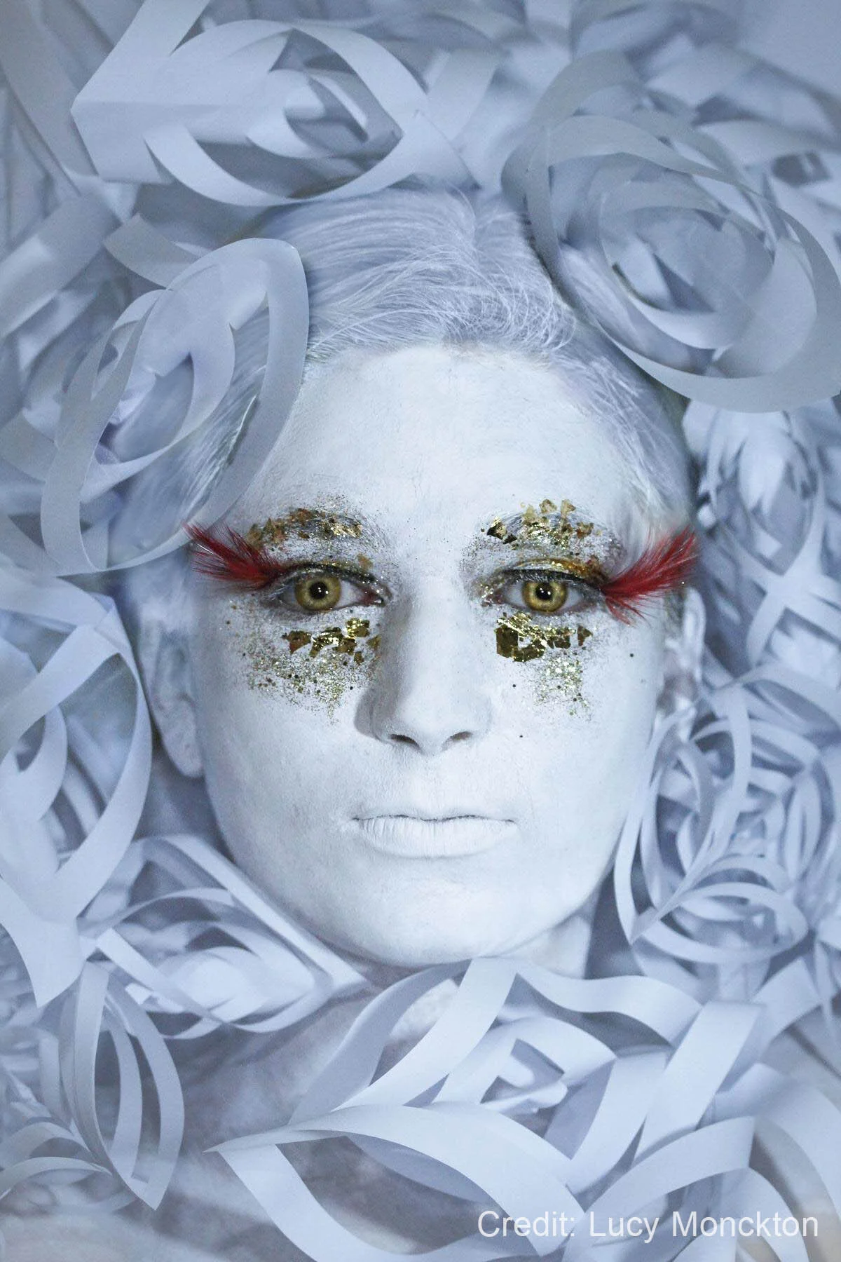 Artistic portrait of a person with white painted skin, surrounded by curled white paper. They have gold decorations around their eyes and red feather-like eyelashes.