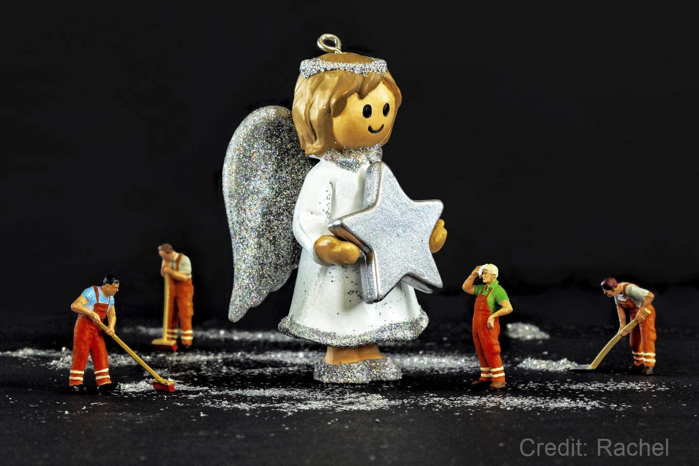 A small angel figurine holding a silver star and surrounded by miniature toy workers cleaning up glitter on a black background.