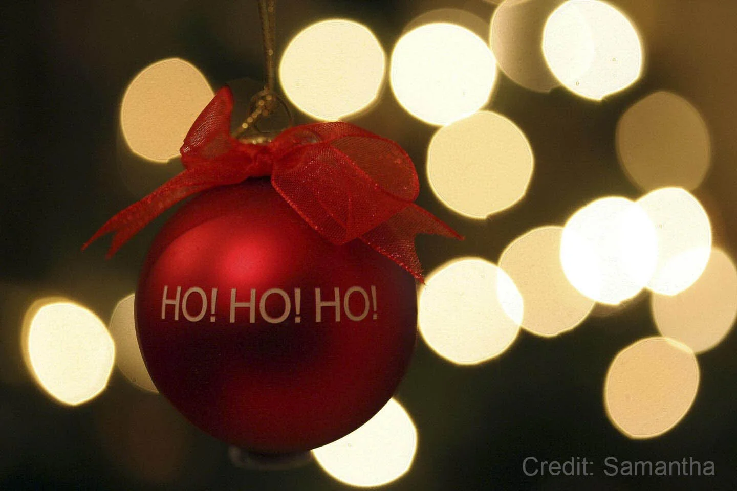Red Christmas ornament with "Ho! Ho! Ho!" text and bow, with blurred lights in the background.