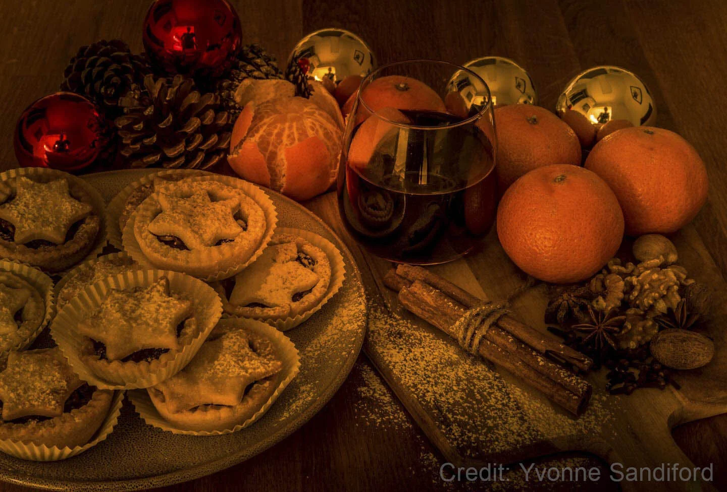 Festive holiday arrangement with mince pies, oranges, cinnamon sticks, pine cones, red and gold ornaments, a glass of wine, walnuts, and star anise on a wooden table.