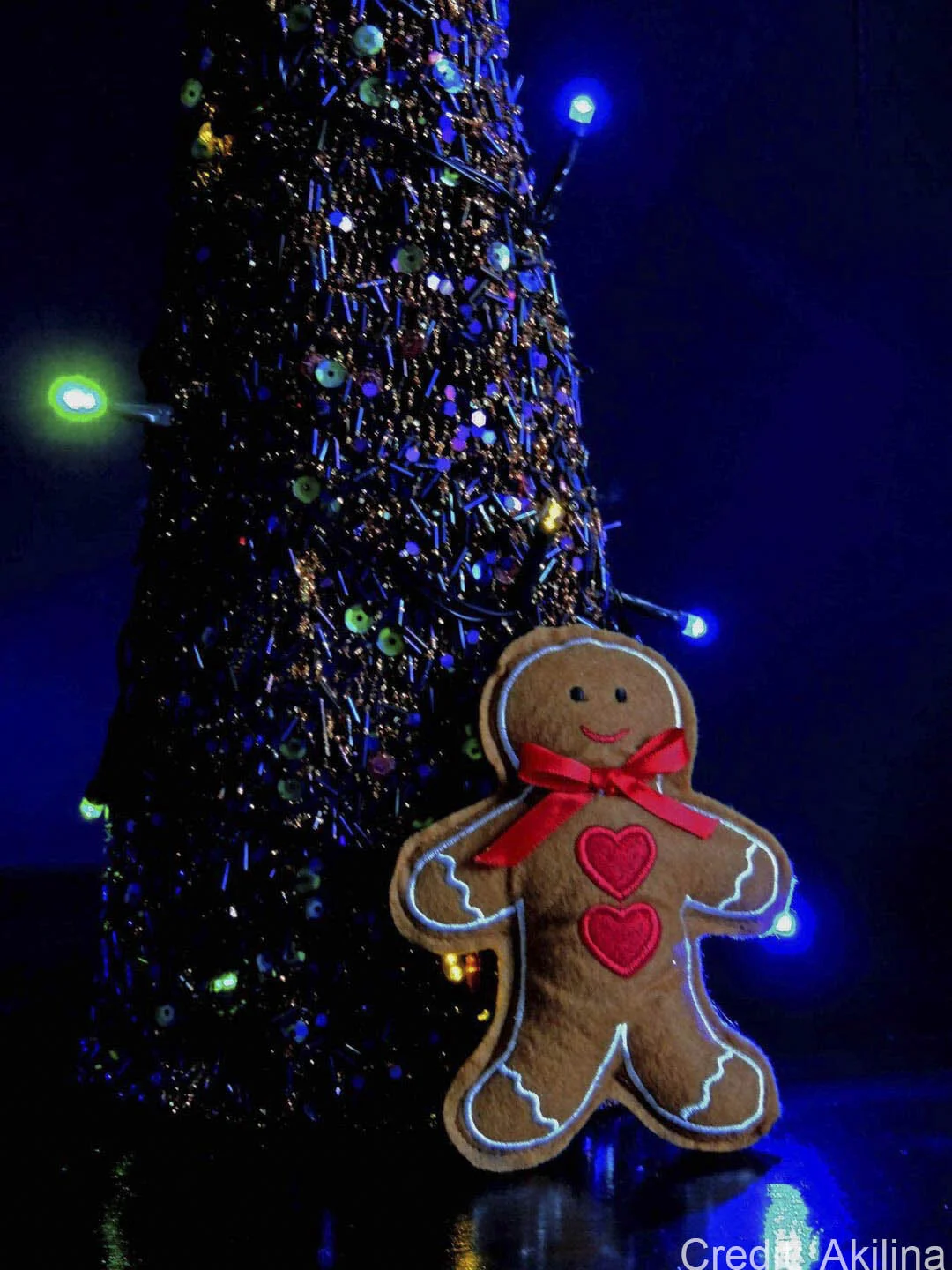 A decorative gingerbread man plush with a red bow and heart shapes stands in front of a sparkling, dark-colored Christmas tree adorned with blue and green LED lights.