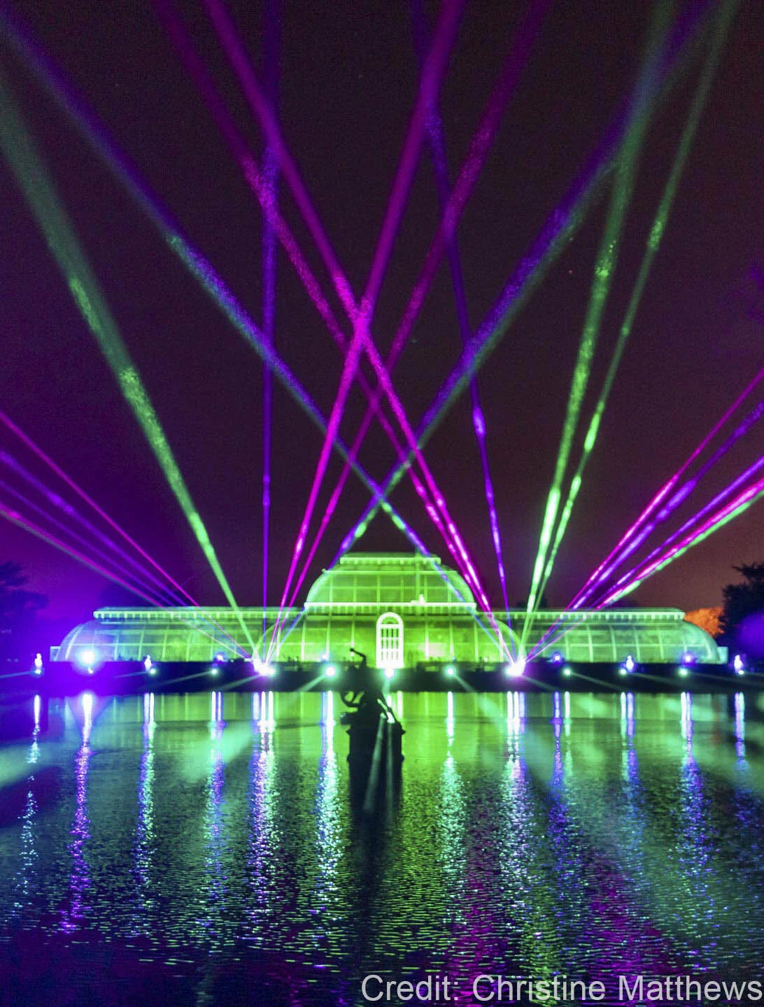 Colorful laser light show illuminating a glasshouse at night with reflections on a water surface, creating a vibrant and dynamic atmosphere.