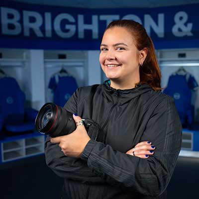 Amy Moore smiling and holding a Canon camera in a Brighton & Hove Albion locker room, with “BRIGHTON &” visible on the wall behind her. She wears a black jacket and stands with arms crossed, conveying confidence and professionalism.