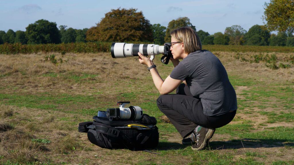 Connie Kinley kneels in an open field, shooting with the Sony A1 II and FE 200–600mm G OSS, camera bag and spare gear on the ground beside her.