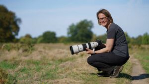 Connie Kinley smiles at the camera while holding a Sony A1 II with FE 200–600mm G OSS, resting in a grassy landscape.