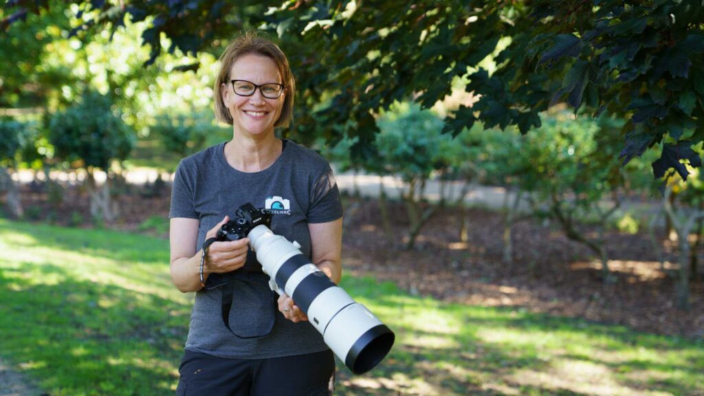 Portrait of Connie Kinley standing under trees, holding a Sony A1 II and FE 200–600mm G OSS, smiling in soft natural light.