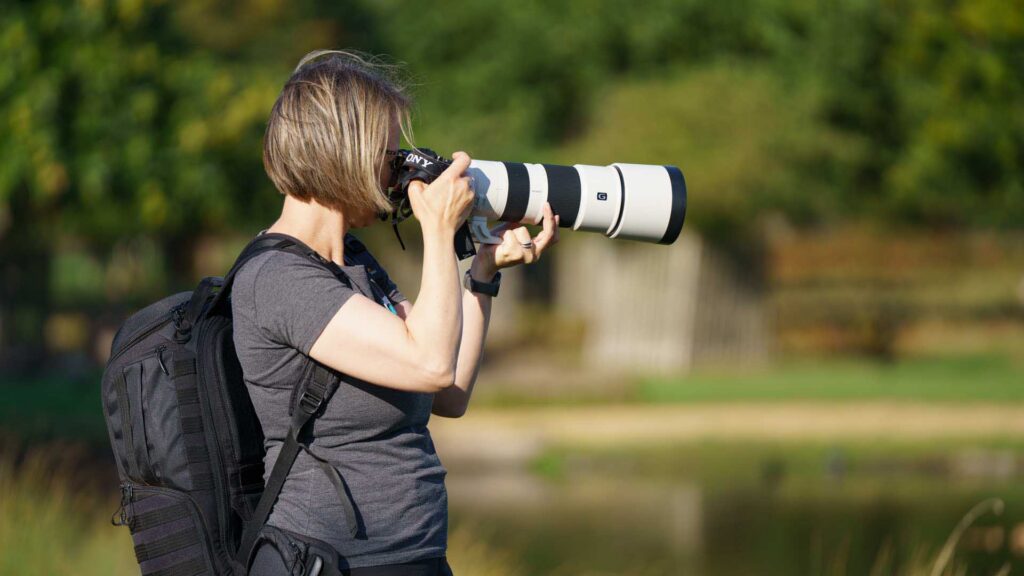 Side view of Connie Kinley standing and photographing with a Sony A1 II and FE 200–600mm G OSS, backpack on, against a blurred park background.