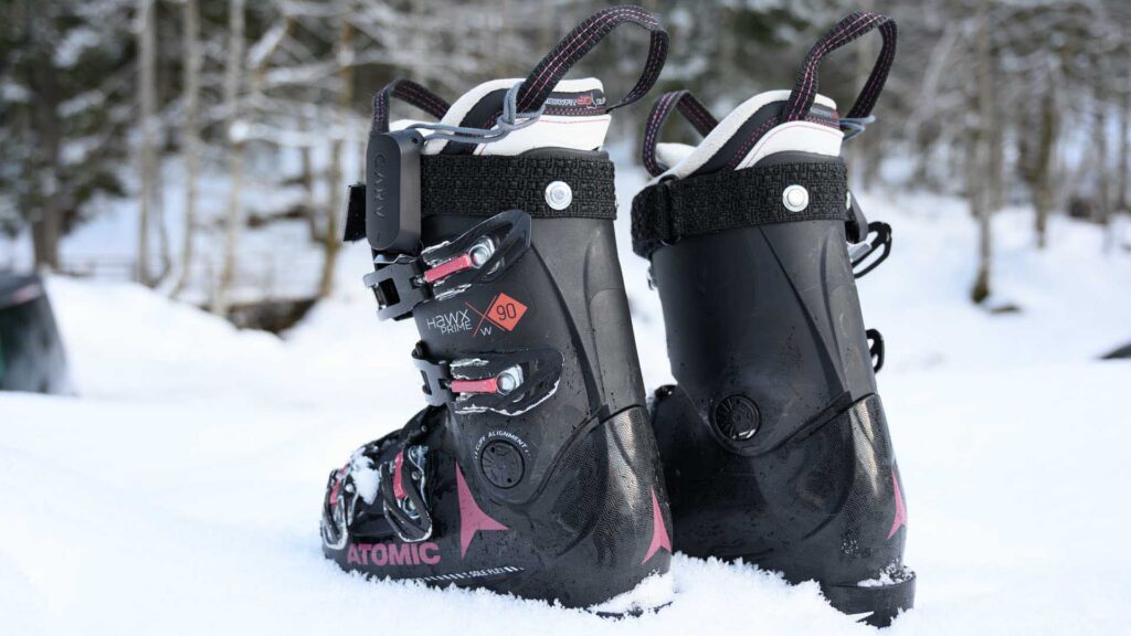 Pair of black Atomic Hawx Prime 90 W ski boots standing in the snow, with Carv sensor mounted on the upper cuff.
