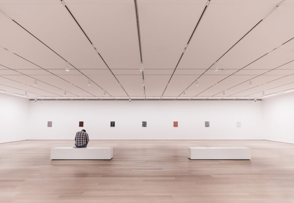 Lone visitor sits on a white bench in a vast, minimalist gallery, small artworks spaced along a blank wall under a grid of ceiling lights.