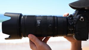 Nikon NIKKOR Z 70-200mm f/2.8 VR S lens mounted on a Nikon Z-series camera, held in both hands at the beach with water droplets on the barrel and the ocean blurred in the background.
