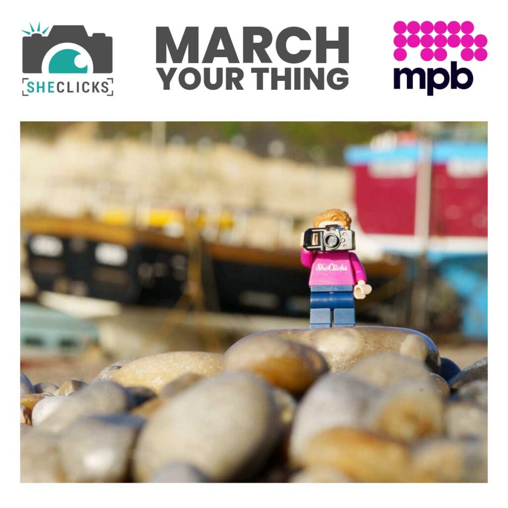 Banner with SHECLICKS logo, bold text “MARCH YOUR THING,” and mpb logo above a photo of a Lego mini figure wearing a pink “SheClicks” t-shirt and holding a camera, standing on pebbles with blurred boats in the background.