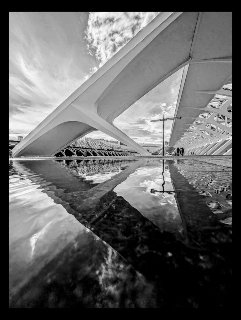 Black and white wide-angle view of sweeping white architectural arches reflected in a shallow pool, with a street sign at centre and a few small figures walking beneath the structure.