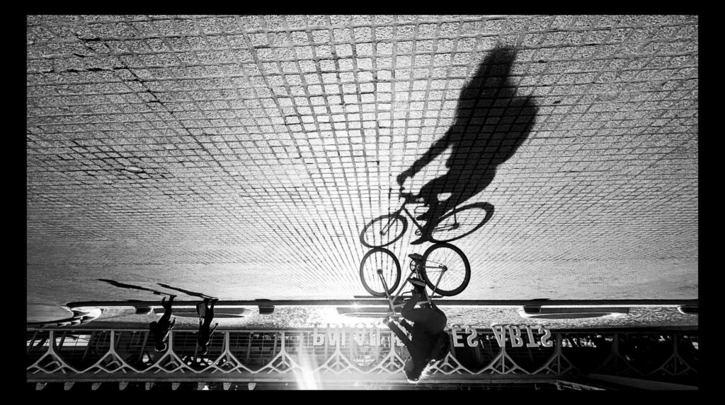 Upside-down black and white scene of a cyclist and their elongated shadow crossing a tiled plaza, with the sun flaring near a modern building façade and reversed signage visible in the background.