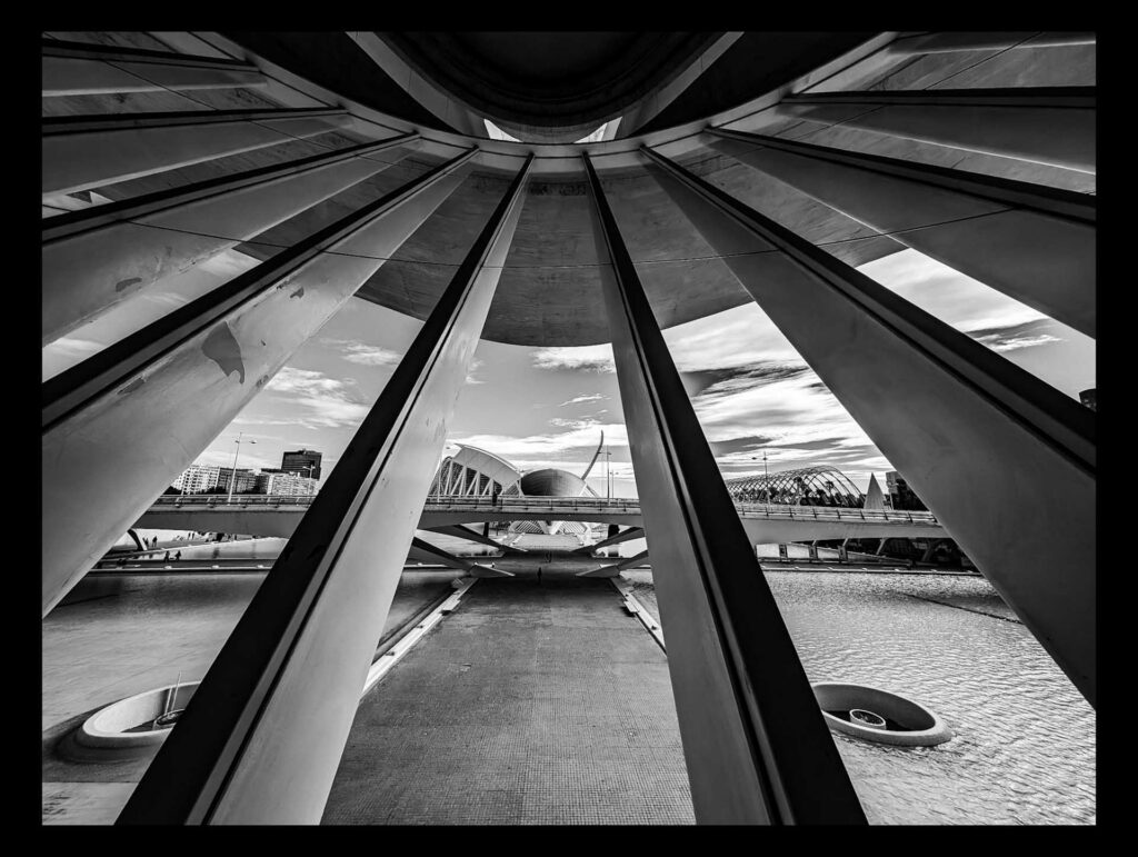 Black and white view framed between massive angled beams, looking toward a futuristic bridge and domed structures over water, with a city skyline in the distance.