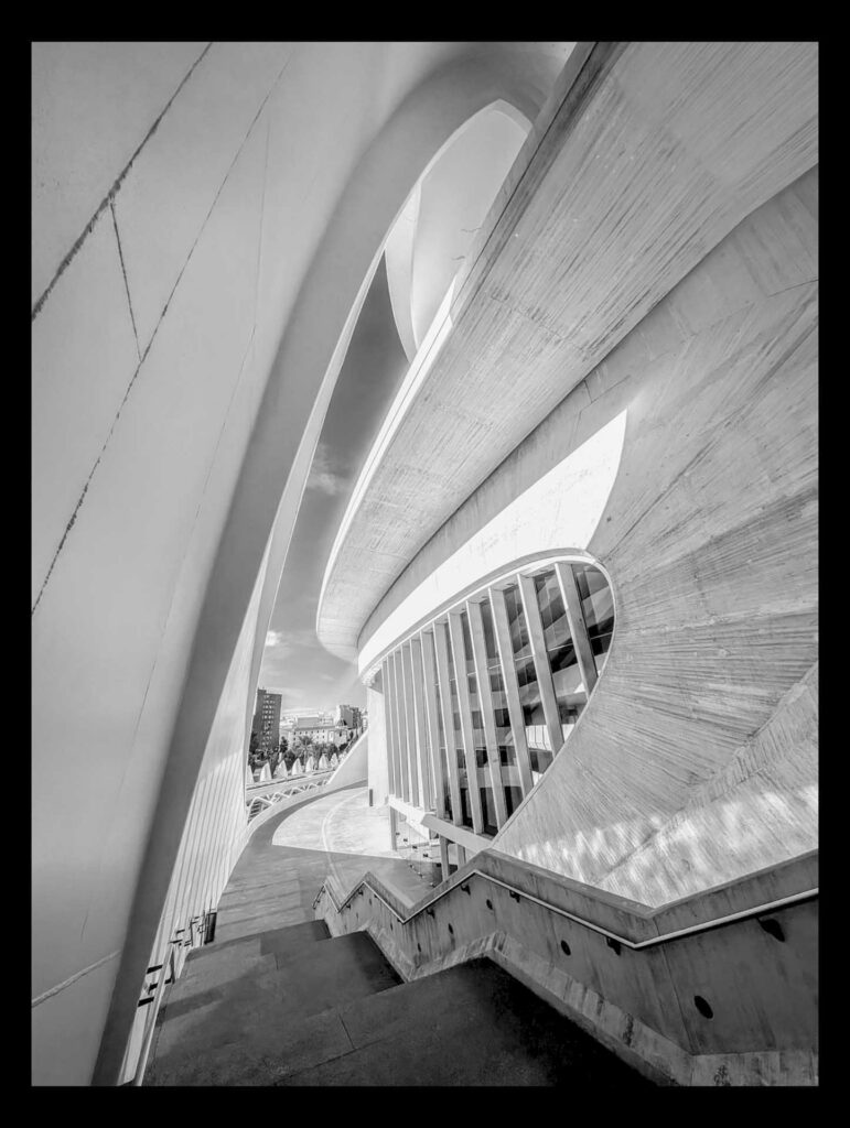 Black and white architectural composition of curved concrete forms and a stairway descending beside a ribbed, oval windowed façade, leading toward an open plaza.