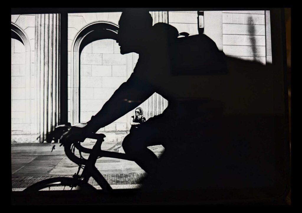 High-contrast black and white silhouette of a cyclist riding past a classical stone building with arched windows, while a seated person in sunglasses is visible in the bright background.