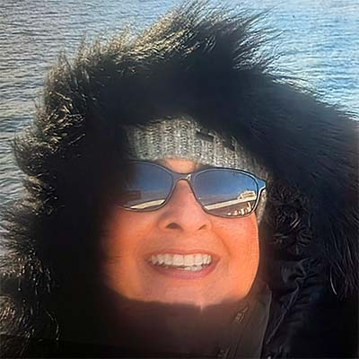 Smiling woman wearing dark sunglasses and a fur-lined hood over a knit headband, with sunlight reflecting on water in the background.