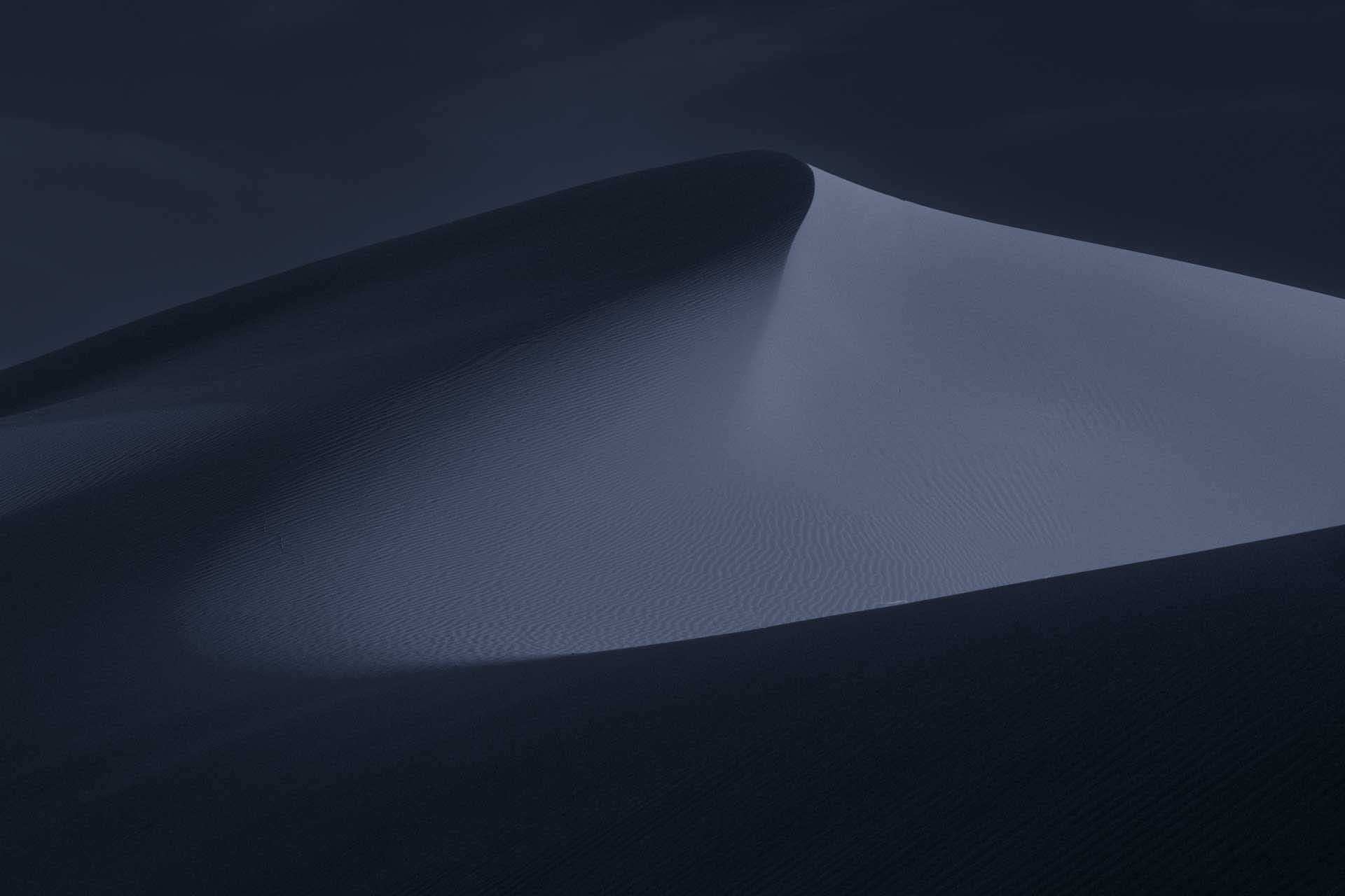 Minimalist blue-toned sand dune with a smooth crest and delicate wind-shaped ripples under a dark twilight sky.